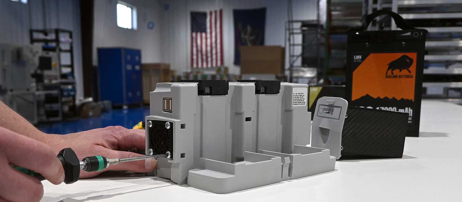 A battery charger is being assembled while fully assembled battery packs are positioned behind the charger.